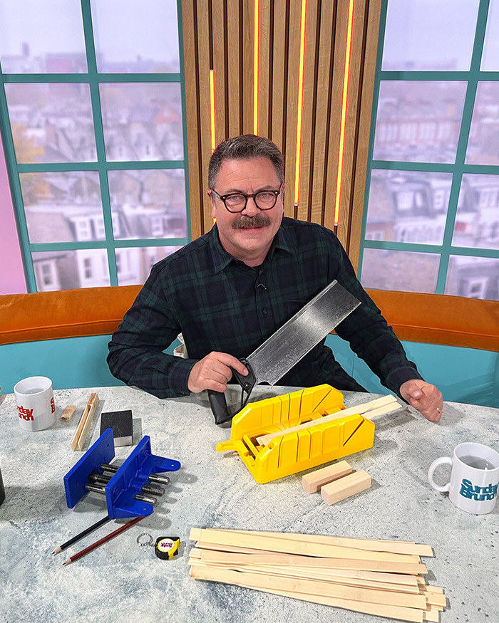 Man with mustache and glasses holding a saw on a talk show set amid woodworking tools during live broadcast incident.