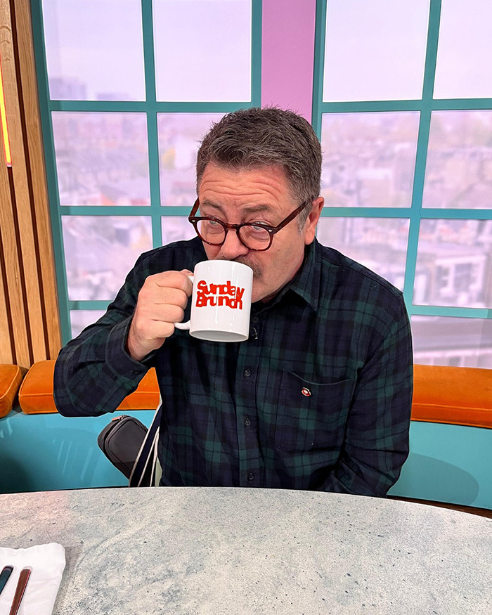 Man wearing glasses drinks from Sunday Brunch mug during live broadcast amid Nick Offerman's x-rated comment incident.