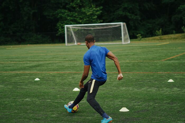Man wearing blue shirt practicing soccer on a green field with goalposts, illustrating positive stereotypes about countries.