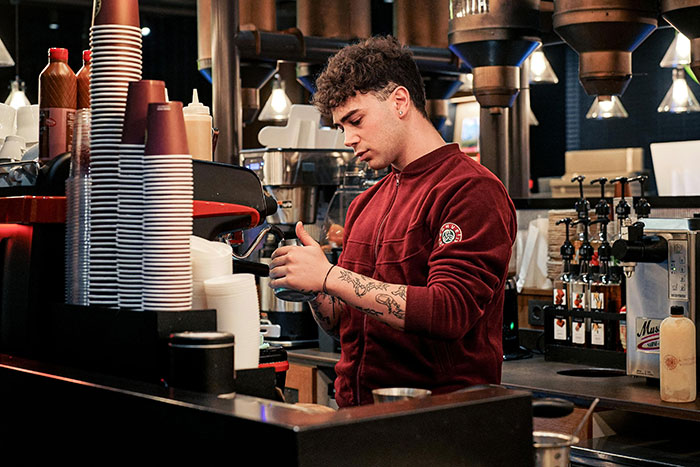 Gas station worker in a red jacket preparing coffee behind the counter with cups and syrup bottles in the background