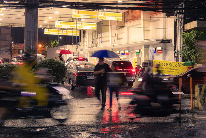 People with umbrellas walk past a busy gas station at night with cars and motorcycles in rainy conditions.