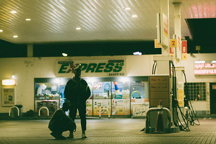 Two gas station workers at night near fuel pumps outside a brightly lit convenience store sharing their wildest stories.