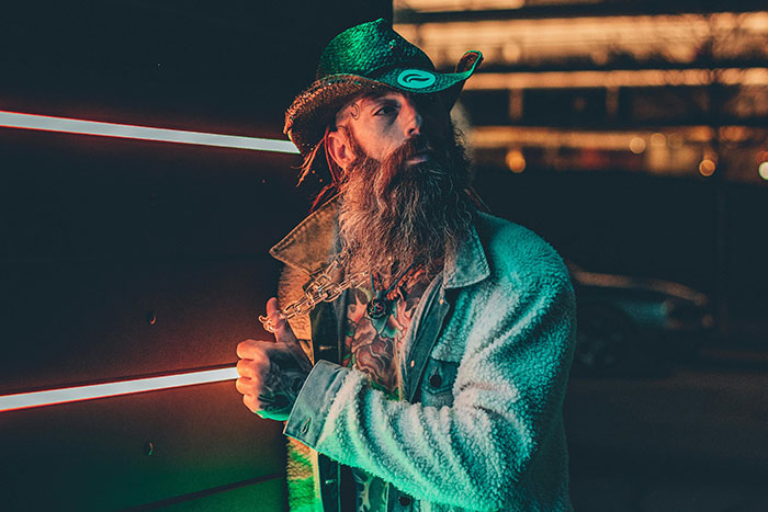 Bearded man in cowboy hat and jacket standing near neon-lit wall, illustrating gas station workers sharing wild stories.