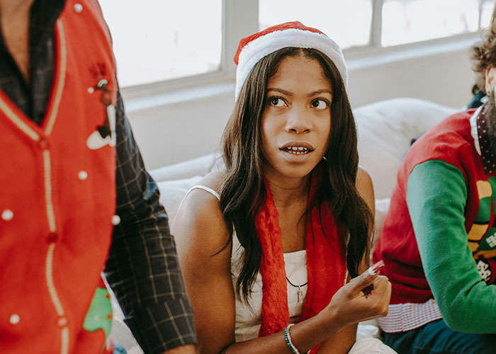Woman in a Santa hat looking confused during family Christmas traditions with others wearing festive sweaters nearby. Woman in a Santa hat looking confused during family Christmas traditions with others wearing festive sweaters nearby.