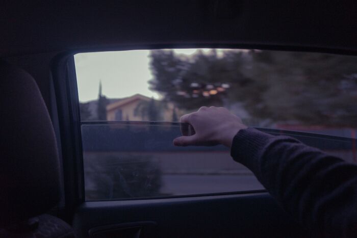 View from inside a vehicle with a person’s hand resting near the window, capturing a quiet road scene at dusk.