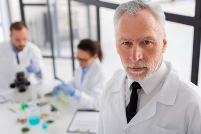 Serious senior man in lab coat with colleagues working in the background, representing boss who insists on strict by the book rules.