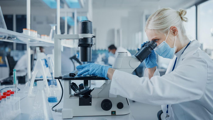 Scientist in lab coat and mask using microscope in a busy laboratory illustrating strict office rules by the book.