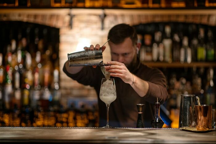 Bartender pouring a cocktail into a glass with ice in a dimly lit bar, illustrating food and drink complaints humor.
