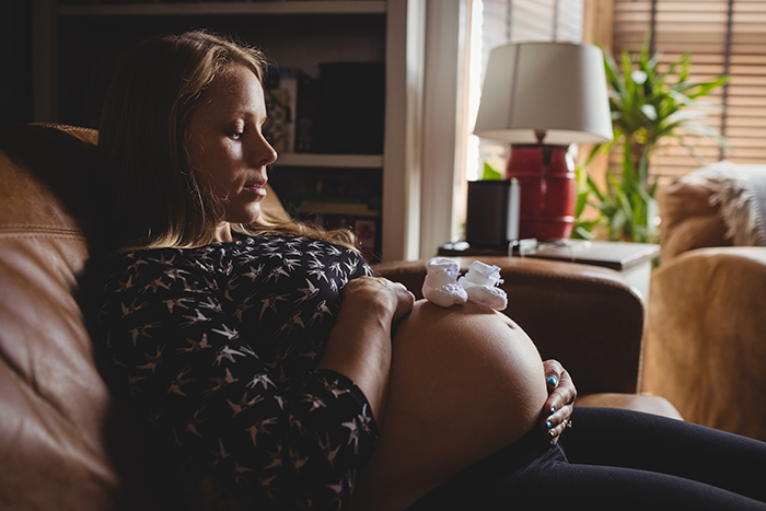 Pregnant woman resting on couch, holding belly with baby booties, symbolizing pregnancy and postpartum struggles.