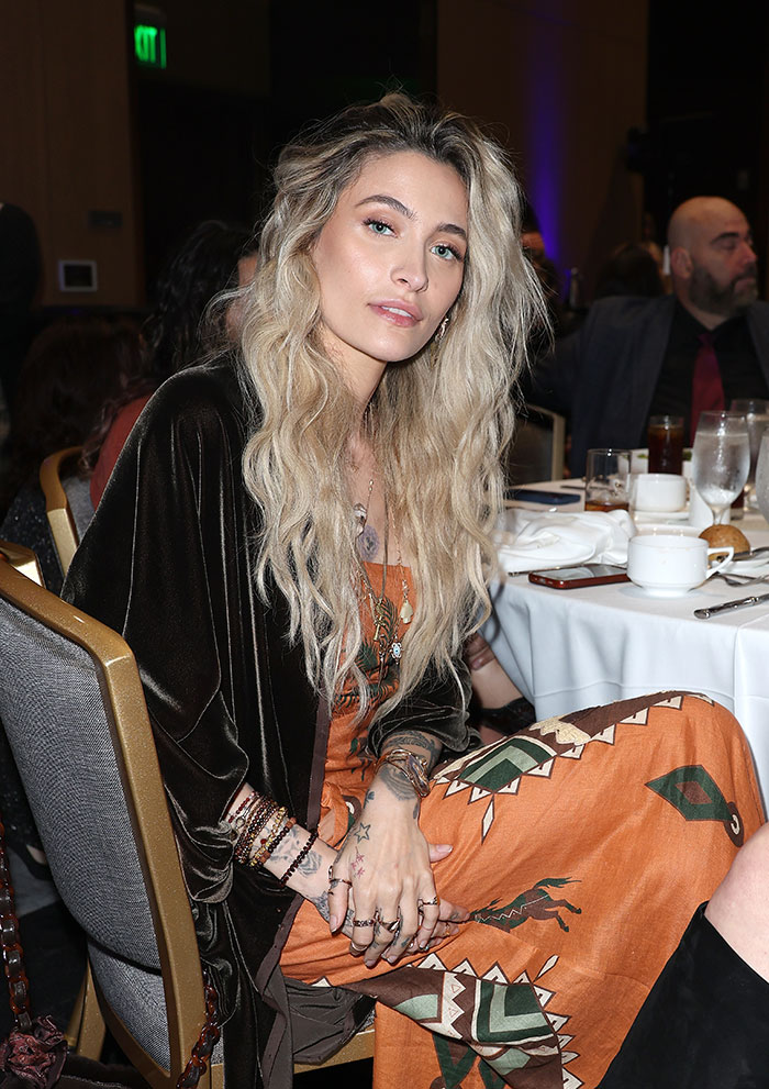 Paris Jackson seated at an event, wearing a patterned dress and showcasing her natural face without visible deformities from substance use.