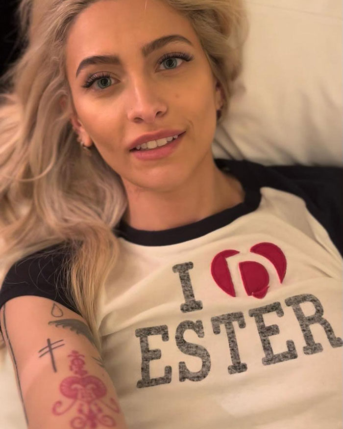 Paris Jackson lying down with tattoos on arm, smiling and wearing a white shirt with black sleeves.