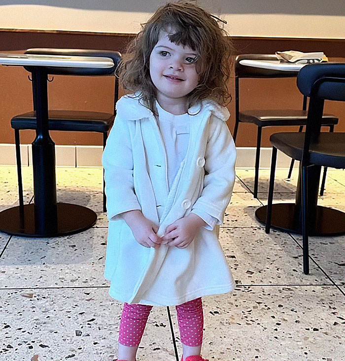 Toddler girl wearing white coat and pink leggings standing indoors near tables and chairs, related to hot car child safety incident.