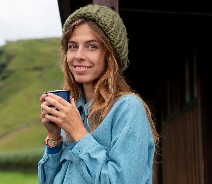Young woman smiling outdoors, wearing a knit hat and blue coat, enjoying a warm drink after walking away from old lives.