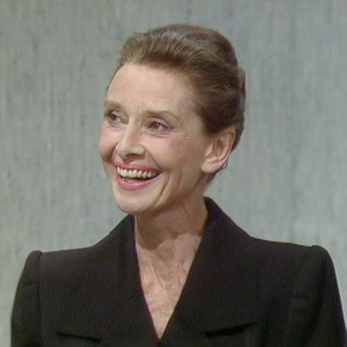 Audrey Hepburn smiling in a black outfit, showing natural aging and wrinkles in a candid moment.