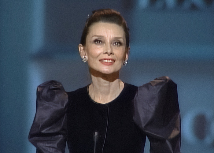 Audrey Hepburn smiling onstage wearing a black dress with puffed sleeves, highlighting reactions to her aging.
