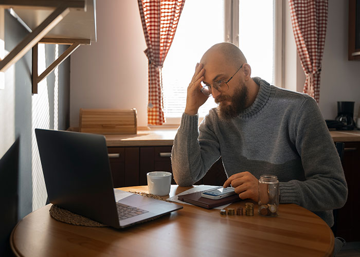 Man looking upset while checking finances at home with laptop and coins on table about discontinued things and foods.