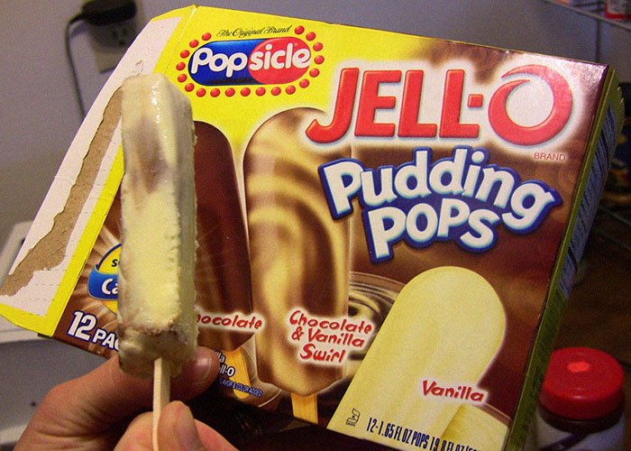 Hand holding a partially eaten Jell-O Pudding Pops ice cream in front of a vintage discontinued frozen treats box.