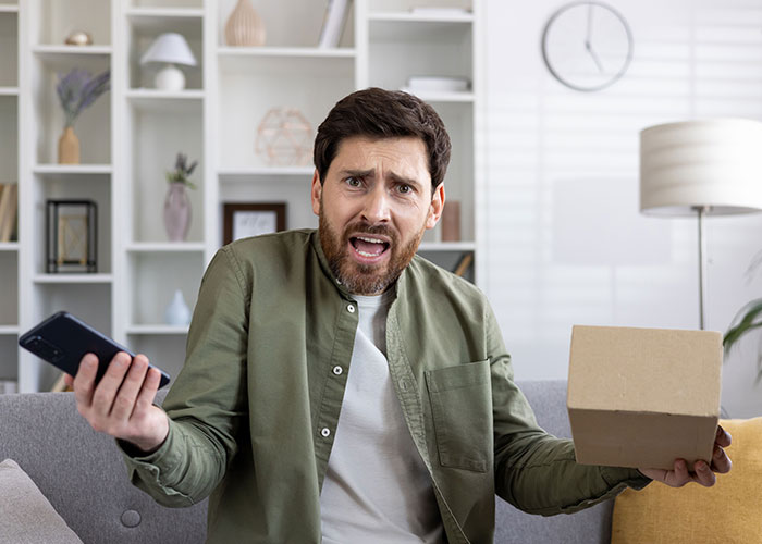 Man frustrated holding a package and phone at home after company sends packages to wrong address by mistake.