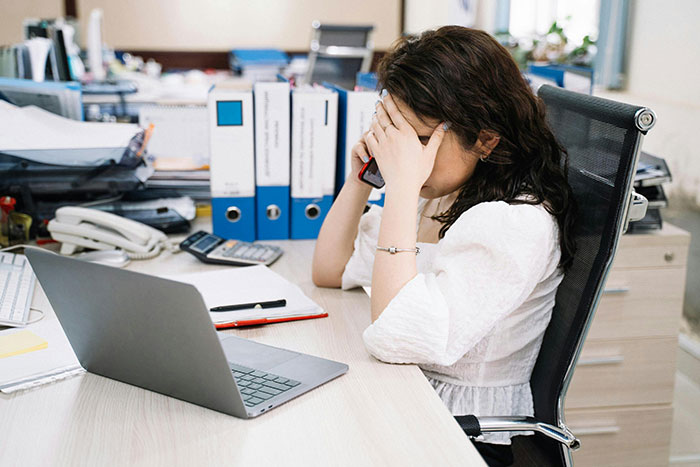 Woman frustrated at office desk, holding phone and covering face, illustrating coworker dispute over expensive jacket.