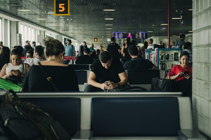 People sitting in an airport waiting area, all looking at their phones, showing relatable everyday behaviors.