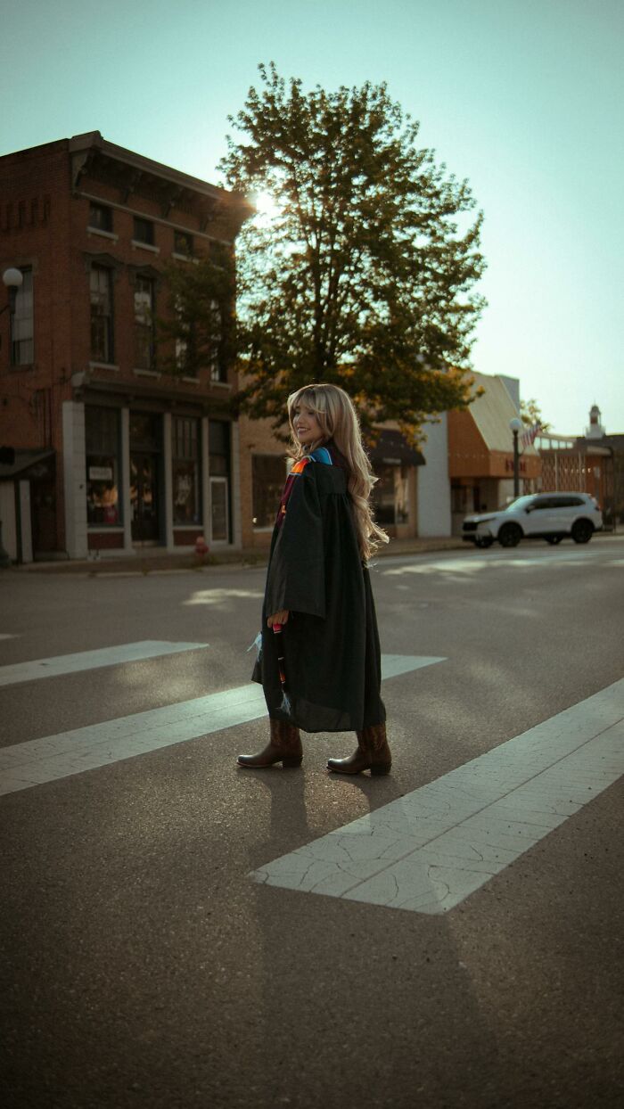 Young woman in a graduation gown standing on a street crosswalk during sunset capturing bizarre moments wonder.