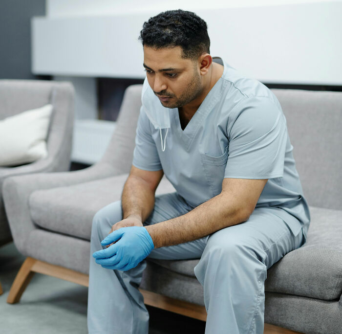Male doctor in scrubs sitting on a couch looking thoughtful, reflecting on eerie patient encounters that haunt doctors.