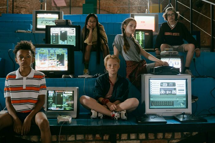 Group of kids with vintage computers and game consoles playing classic 80s and 90s references together indoors.