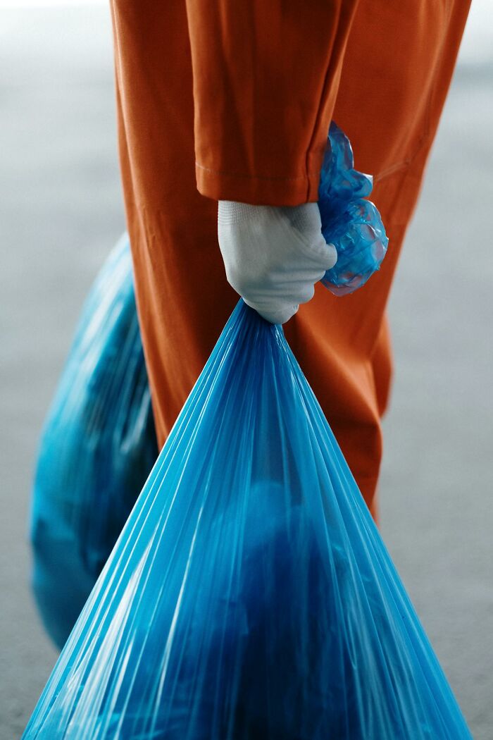Garbage man in orange uniform holding a tied blue trash bag while on the job collecting waste outdoors.
