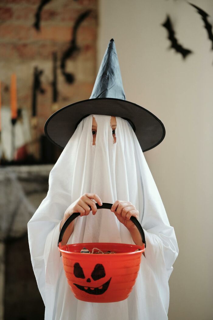 Child in homemade ghost costume with witch hat holding a Halloween bucket, reflecting childhood habits and memories.