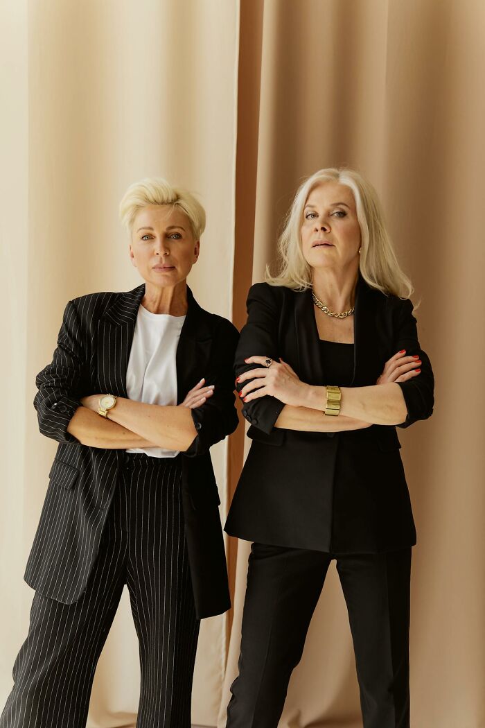 Two confident older women with arms crossed wearing black suits, embodying a bold Karen attitude in a neutral setting.