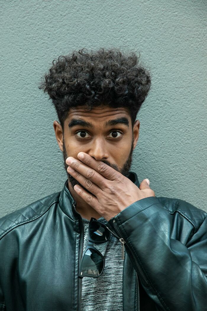 Young man with curly hair covering his mouth in surprise, illustrating funny and serious moments of raising monsters.