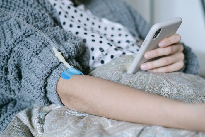 Person in cozy sweater lying in bed with IV drip, holding smartphone, illustrating relatable everyday situations.