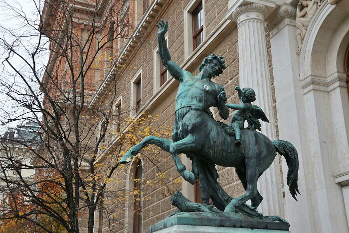Bronze mythology statue of centaur and child near classical building columns in outdoor urban setting.