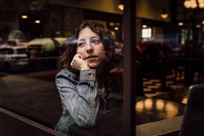 Young woman in a denim jacket looking thoughtfully through a cafe window, reflecting on comically absurd facts.