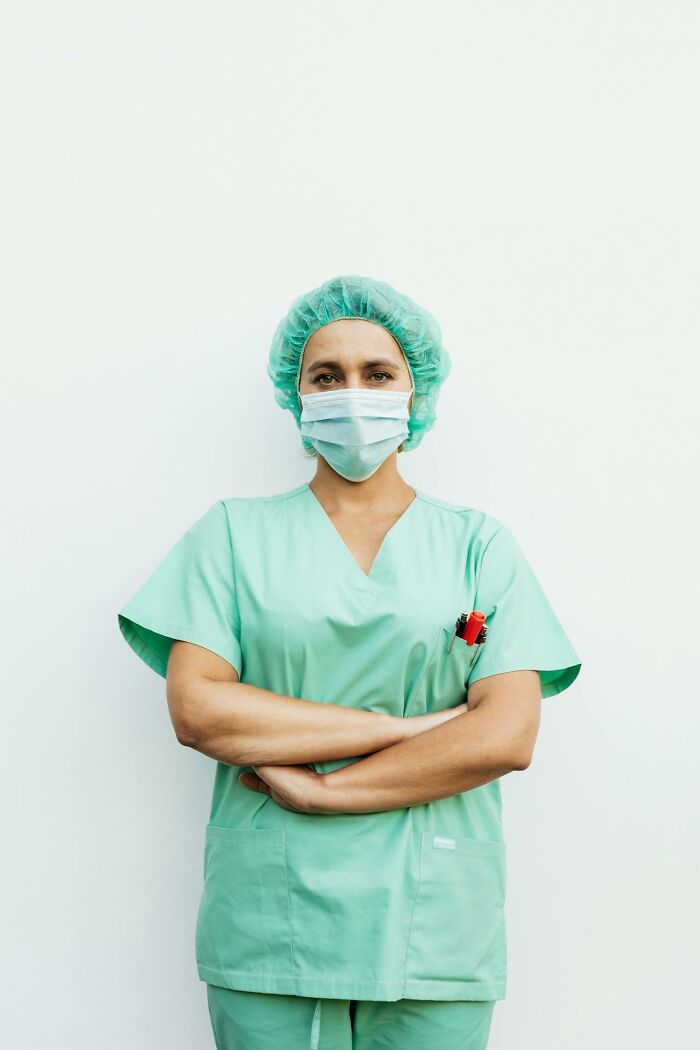 Female healthcare worker in green scrubs and mask standing against white wall with arms crossed, serious expression, Karen example concept.