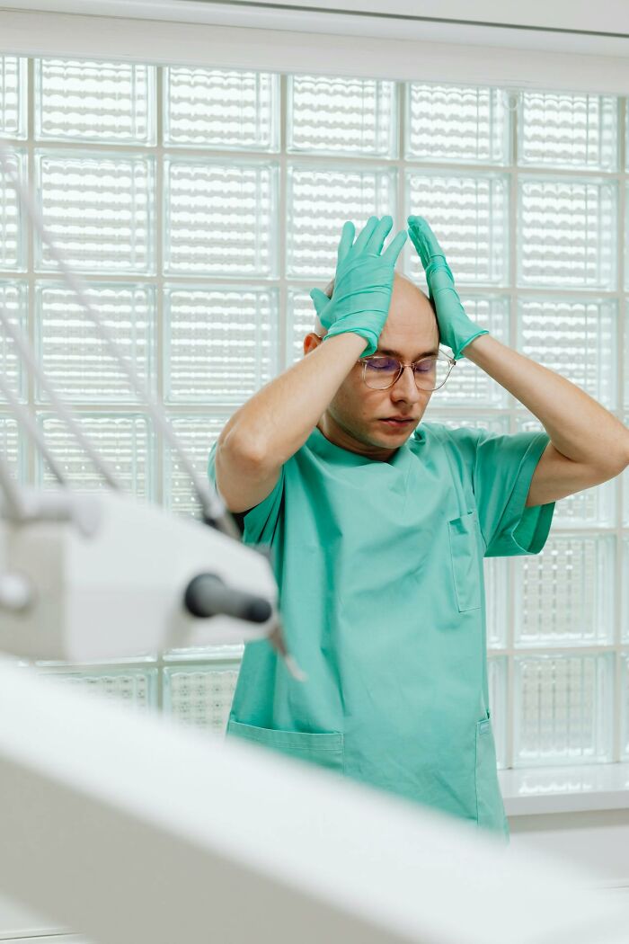 Nurse in green scrubs and gloves looking stressed inside a bright psych ward environment with glass block windows.
