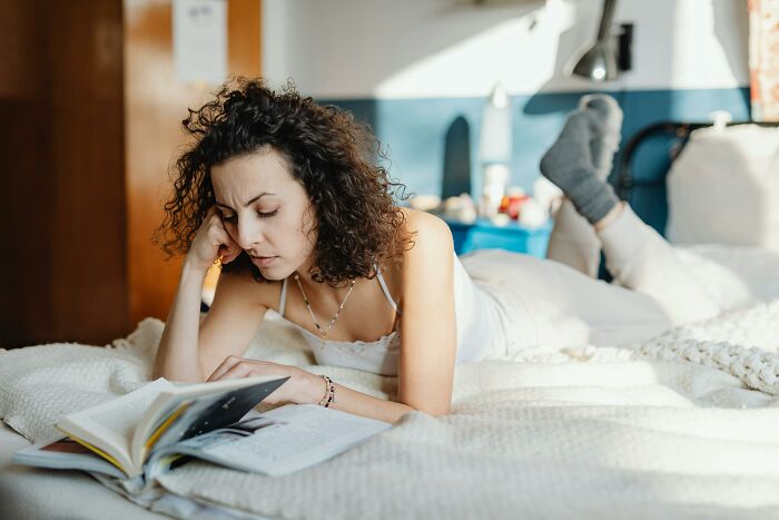 Woman lying on bed reading a book, showing a relatable moment of doing ridiculous but common everyday things.