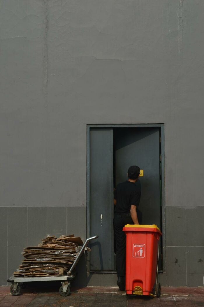 Garbage man opening door next to red trash bin and cart loaded with flattened cardboard on a job site.