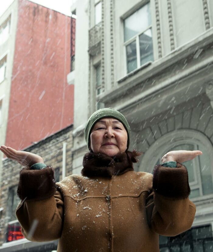 Older woman in winter coat and hat standing in falling snow outside historic buildings, representing nurses of psych wards.