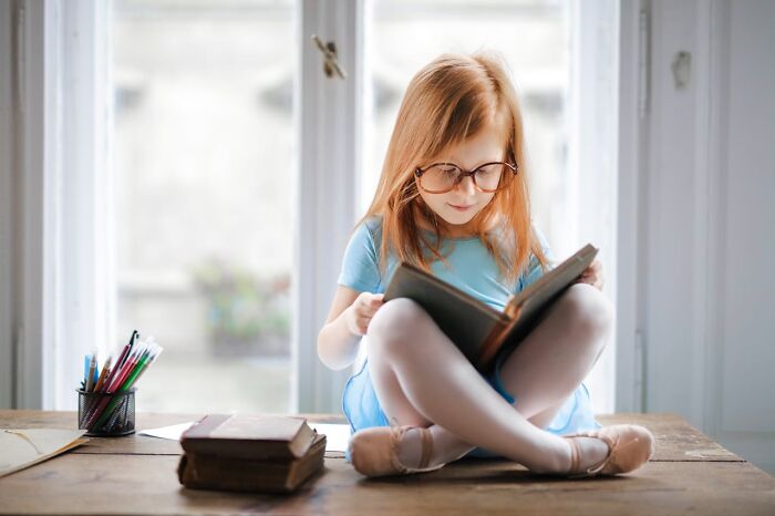 Young girl wearing glasses sitting cross-legged reading a book near window, reflecting childhood habits not normal behavior.