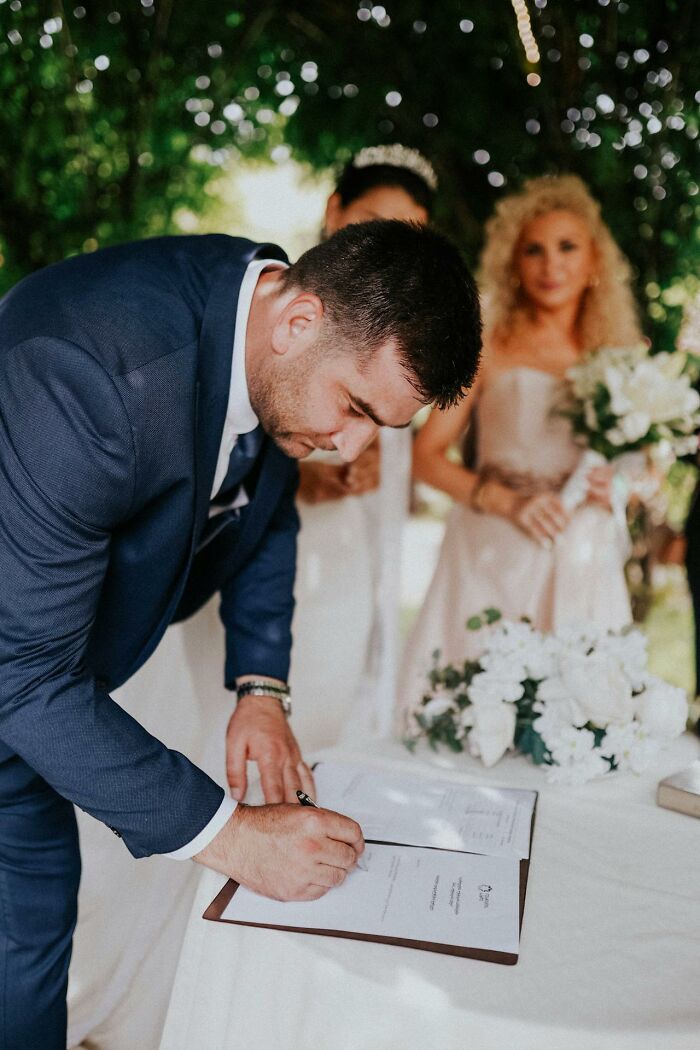 Groom in a blue suit signing marriage documents with bride and bridesmaid holding bouquet in background outdoors.
