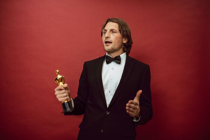 Man in tuxedo holding an award, expressing emotion against a red background, relatable to ridiculous but relatable things.