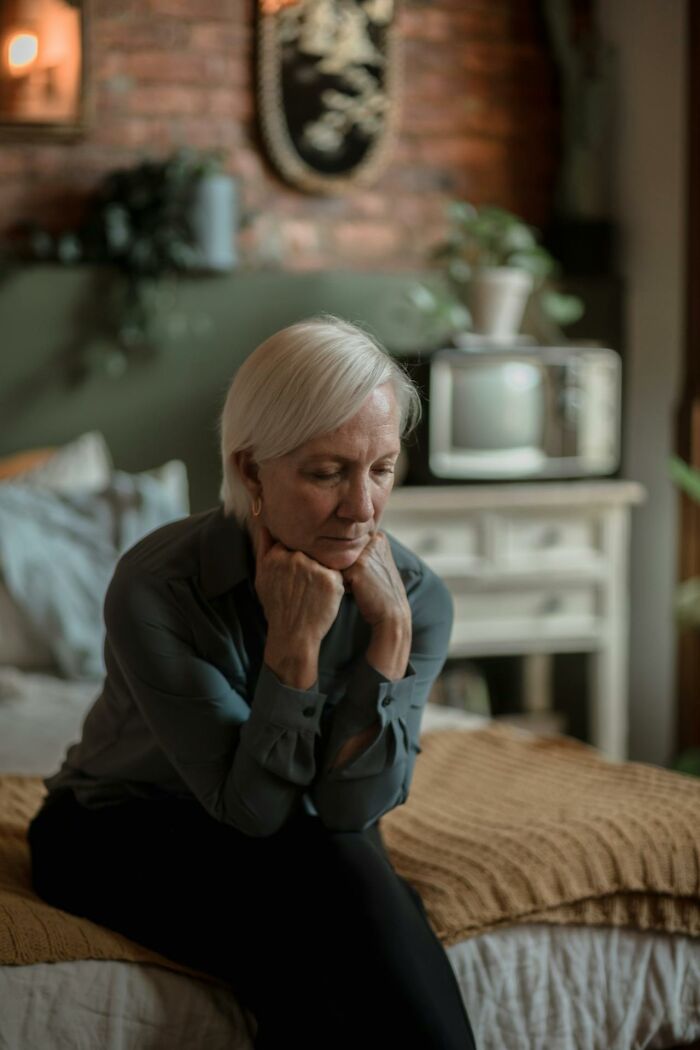 Older woman sitting on bed with hands under chin, appearing thoughtful and reflecting on heavy confessions without charges.