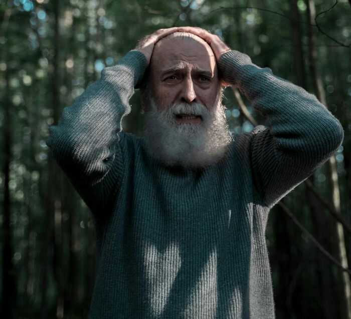 Elderly man with white beard in forest holding head, showing fear and distress, evoking haunting truckers road night stories.