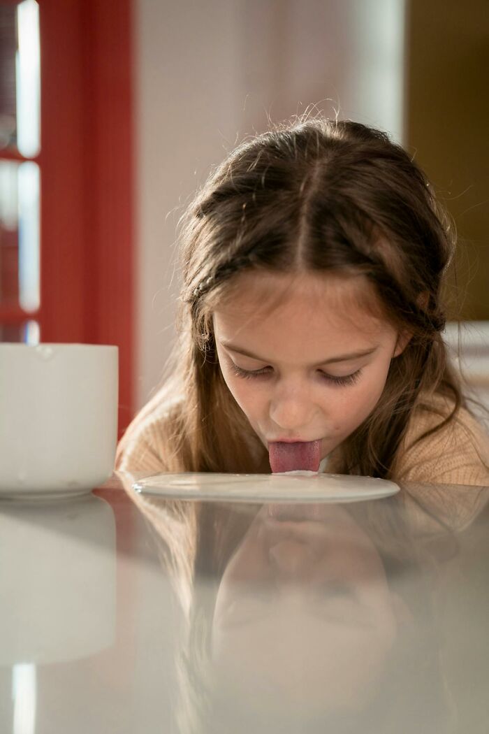 Young girl displaying childhood habits by licking milk spilled on a kitchen table, highlighting unusual childhood behaviors.