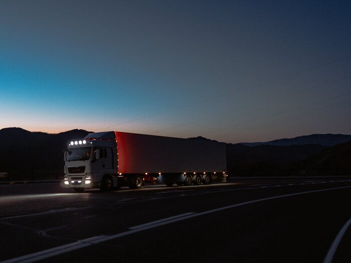 Large semi truck driving on a dark road at night with headlights and mountain silhouette in the background.