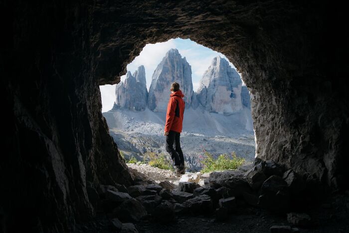 Hiker in a red jacket standing at cave entrance, looking out at rocky mountain peaks, capturing bizarre and creepy nature scene.