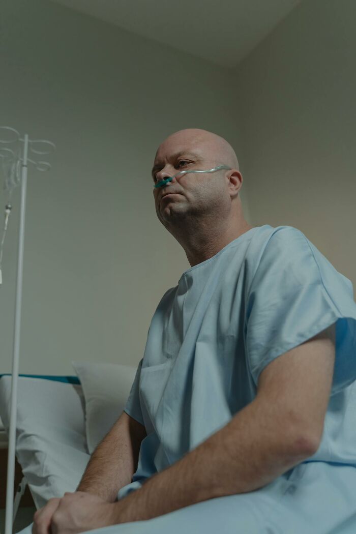 Middle-aged patient in hospital gown with oxygen tube, sitting on bed in a psych ward setting reflecting intense emotions.