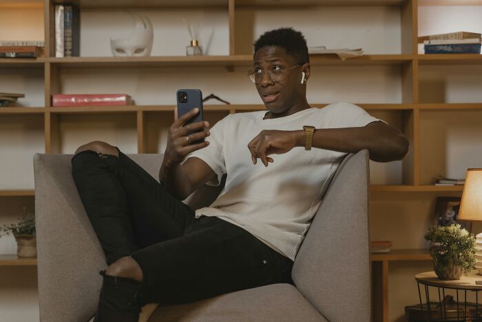 Young man sitting on a chair, wearing earbuds and glasses, engaging with his phone showcasing relatable everyday moments.