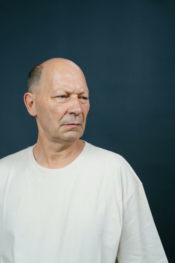 Older man with serious expression in a plain white shirt against dark background, reflecting parents raising monsters moments.